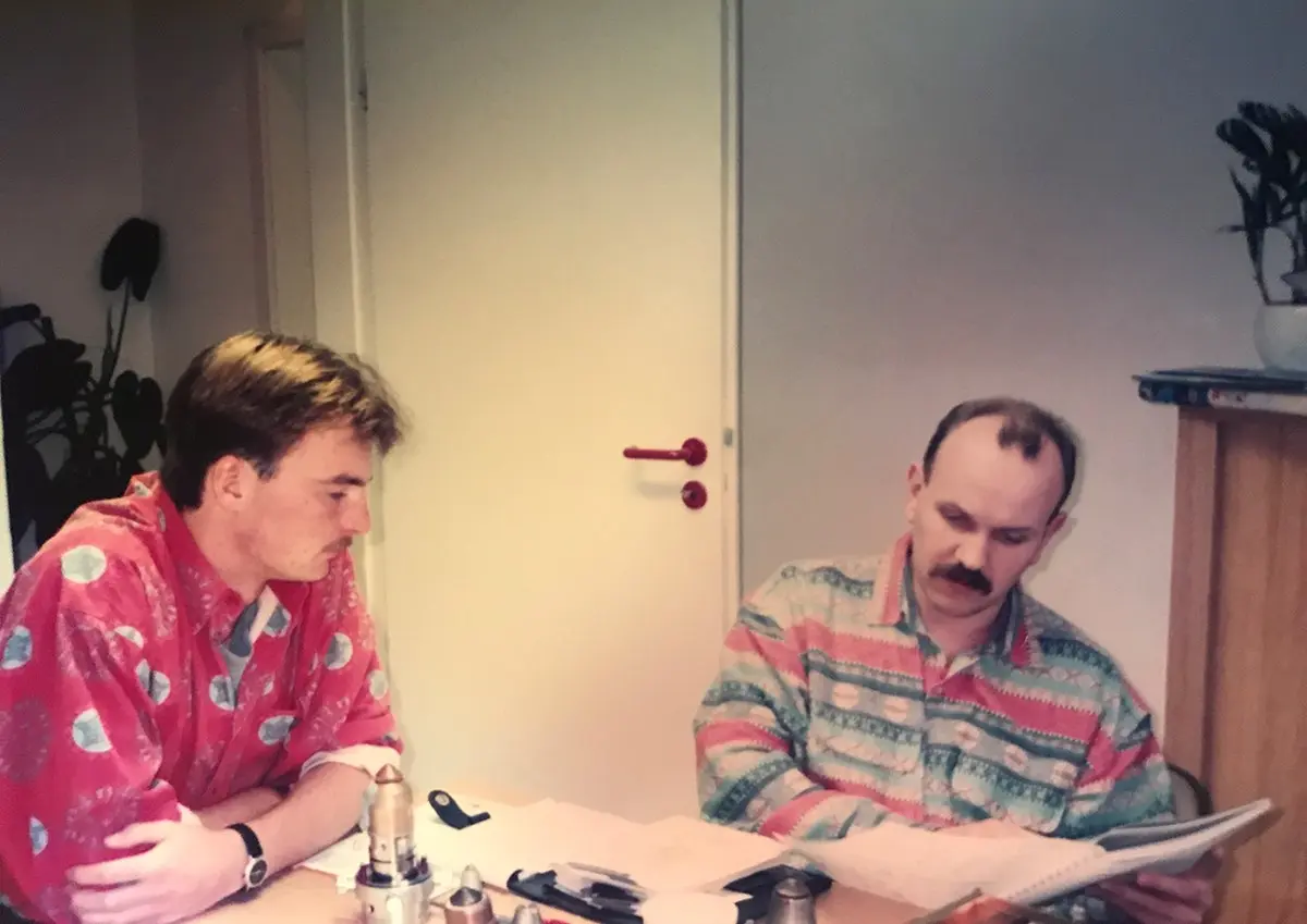 Zwei Männer in bunten Hemden, die an einem Tisch sitzen und Dokumente durchsehen.