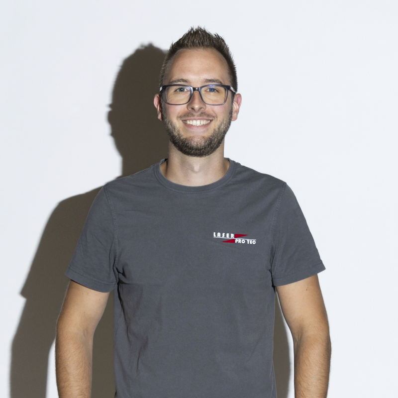 Mann mit Brille und grauem T-Shirt steht vor einem weißen Hintergrund und lächelt.