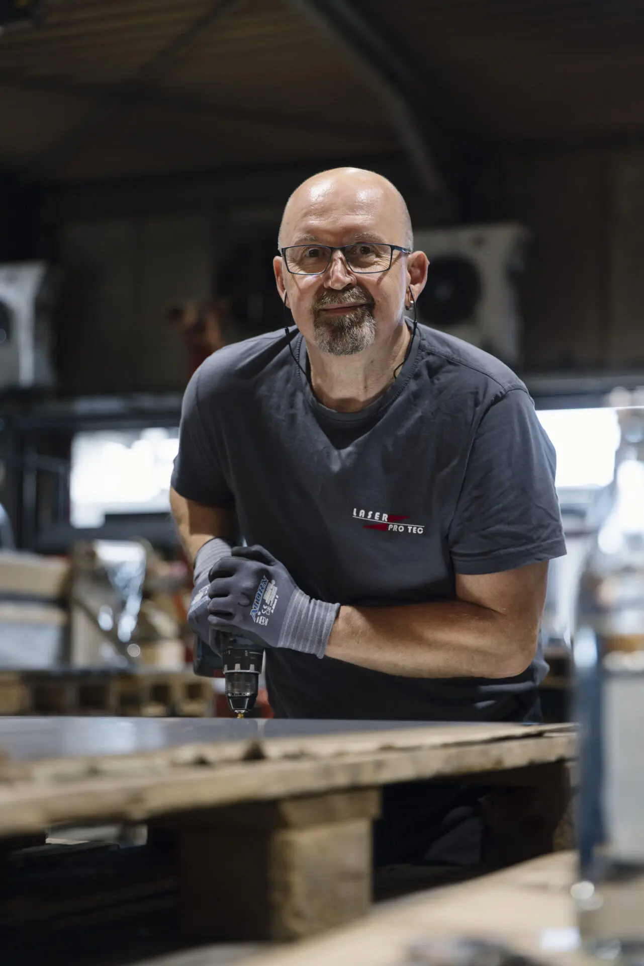 Mann mit Brille und Handschuhen, der eine Bohrmaschine in einer Werkstatt benutzt.